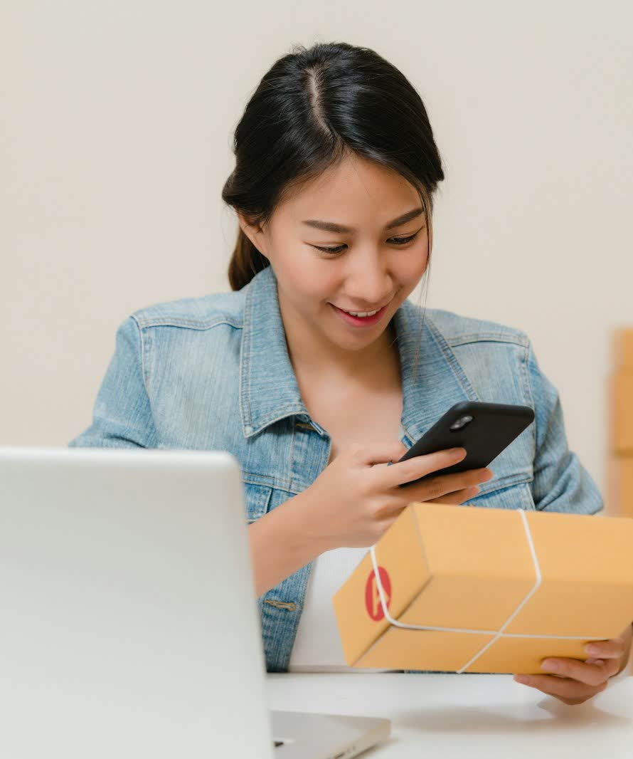 Une commerçante scanne un colis avec son téléphone, à côté de son ordinateur et de cartons d'expédition